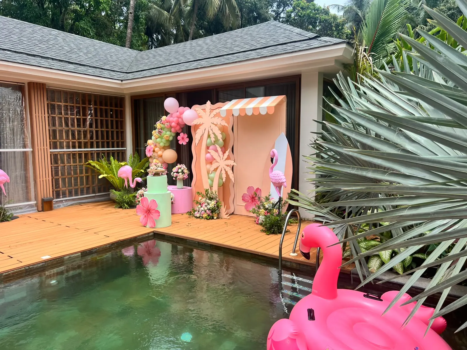 Peach cabana photo booth with palm cut-outs, pink surfboard and flamingo statues by a Goa villa pool by Imagine Decorators