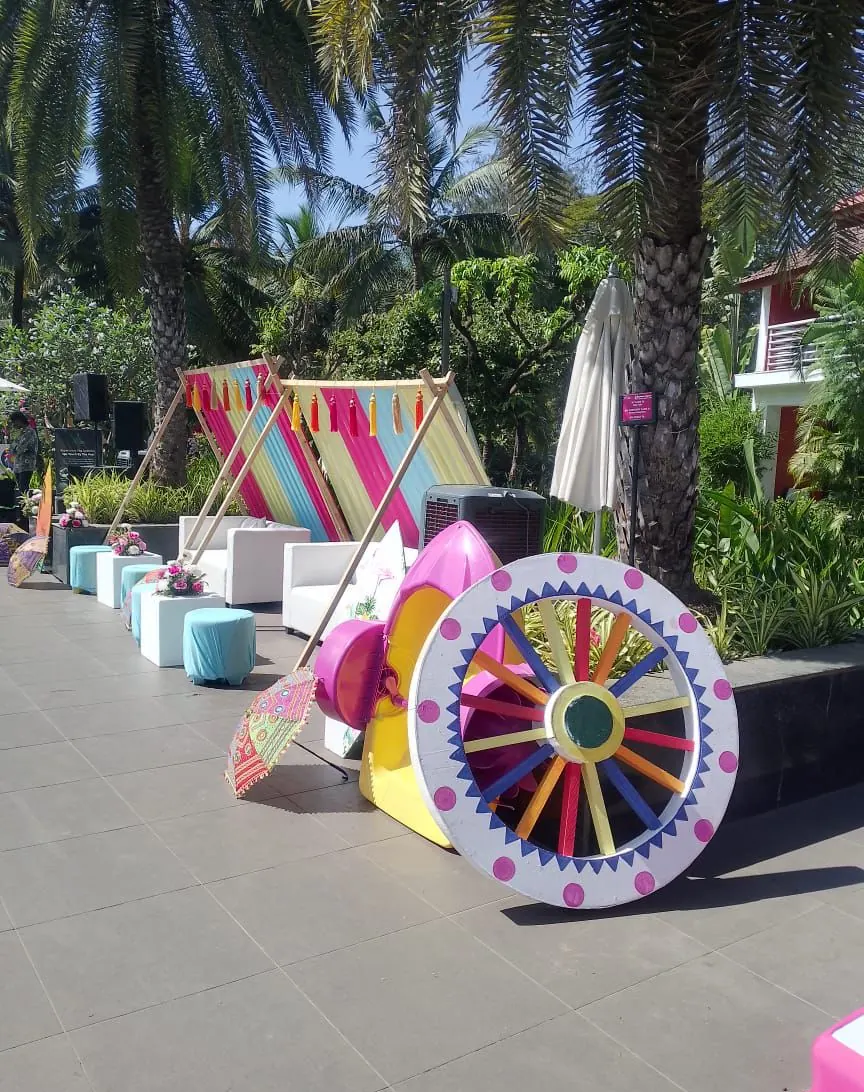 Tassel teepee photo corner with a vintage chariot wheel and pink umbrella props at a Goa haldi by Imagine Decorators