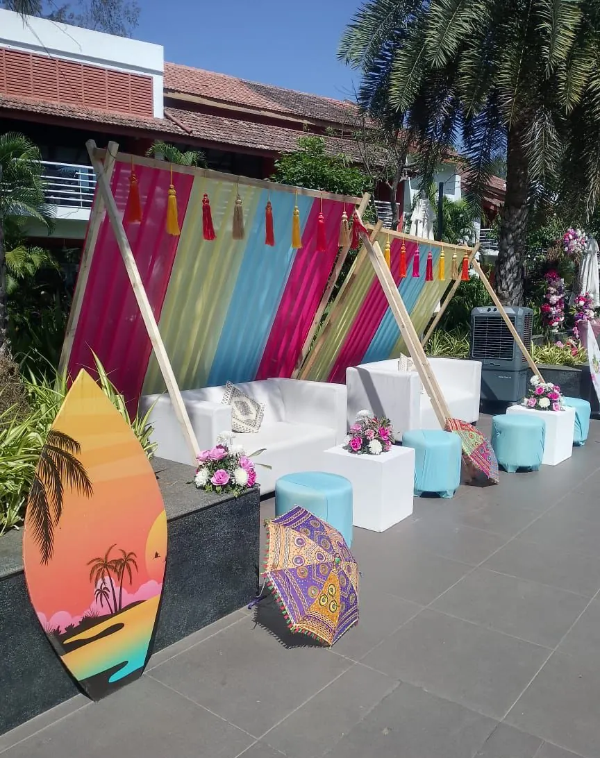 Tassel teepee lounge backdrop with white sofas, pink florals and a surfboard prop at a Goa haldi by Imagine Decorators